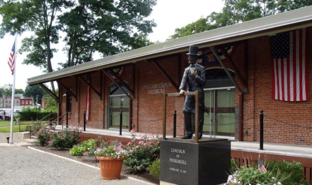 Brick building, original Peekskill train depot. Flowers and statue of Abraham Lincoln in the foreground and two American flags in the background